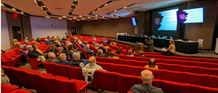 A group of adults are sitting in red theater chairs looking at a presentation on a large screen at the front of the auditorium