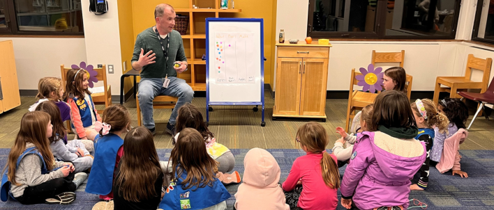 A group of girl scouts are sitting in a circle listening to a male librarian read a story