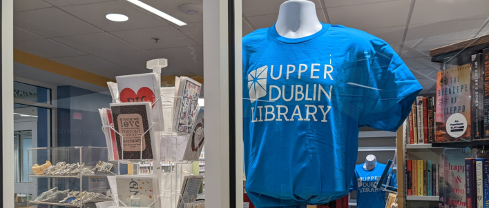 A photo taken from the outside of the friends shop looking in showing a library t-shirt and a rack of greeting cards